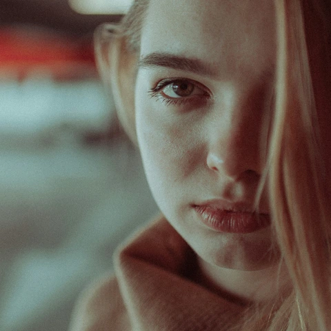 Tightly cropped portrait in muted light: blonde hair covering part of the face, brown coat collar, and a blurred background.