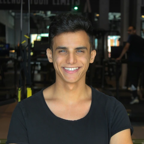 Front-facing portrait with a broad smile: short dark hair and black t-shirt, brightly lit face against a dim indoor background.