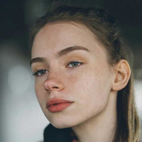 Close-up portrait in soft light: hair pulled back, visible freckles, neutral expression, and gaze directed slightly past the camera.