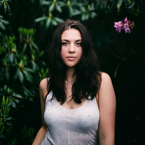 Waist-up garden portrait: long dark hair, light sleeveless top, direct gaze, and dense green foliage with small purple flowers behind.